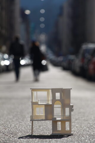 Ein kleines Architekturmodell eines modernen Gebäudes steht auf einer Stadtstraße, im Hintergrund sind verschwommene Figuren und geparkte Autos zu sehen.
