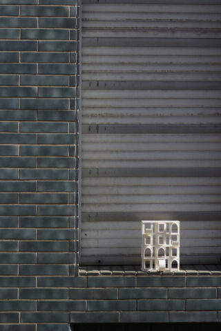 Ein kleines weißes geometrisches Objekt steht auf einem Sims vor einer grün gefliesten Wand und einem geschlossenen Metallladen.