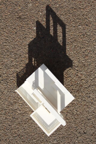 Ein weißes Architekturmodell wirft einen deutlichen Schatten auf eine strukturierte, braune Oberfläche.