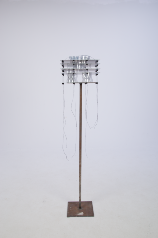 Eine Metallskulptur mit einer zylindrischen, geschichteten Oberseite, von der dünne Drähte herabhängen, montiert auf einer hohen, schlanken Metallstange und einem quadratischen Sockel, vor einem schlichten weißen Hintergrund.