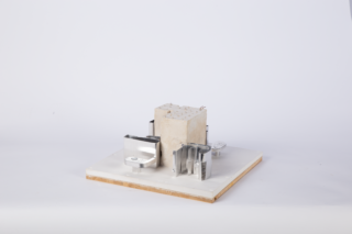 Ein rechteckiger Betonblock mit eingebetteten Metallklammern ist auf einem weißen quadratischen Sockel vor einem schlichten Hintergrund montiert.