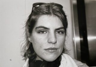 Schwarz-Weiß-Nahaufnahme einer Person mit kurzen Haaren, die eine Sonnenbrille auf dem Kopf trägt und mit neutralem Gesichtsausdruck direkt in die Kamera blickt.