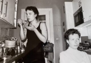 Zwei Personen stehen in einer Küche. Eine steht und isst mit einem Löffel, die andere sitzt an einem Tisch im Vordergrund und schaut weg. Das Foto ist schwarz-weiß.