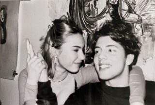 Eine junge Frau und ein Mann sitzen lächelnd eng beieinander. Die Frau hebt den Arm und zeigt mit dem Finger nach oben. An der Wand hinter ihnen ist ein Wandgemälde oder Gemälde zu sehen.