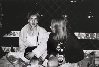 Zwei Personen sitzen nachts auf einer mit Graffiti bedeckten Bank und unterhalten sich. Eine Person blickt in die Kamera, die andere mit einem Getränk in der Hand. Im Hintergrund ist ein Maschendrahtzaun zu sehen.