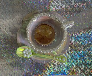 Schillernde Teetasse und Untertasse mit perlenartigen Perlen, einer braunen Flüssigkeit in der Tasse und einem passenden Löffel mit weißen Perlen auf einer glitzernden Oberfläche.