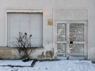 Ein geschlossenes Fenster mit Jalousien und eine Glastür mit gemusterten Bezügen, beide in einer abgenutzten Außenwand eingelassen. Schnee bedeckt den Boden und daneben steht ein blattloser Strauch.