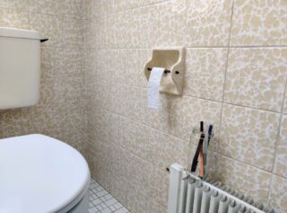 Ein Badezimmer mit gemusterten beigen Fliesen, einer Toilette, einer fast leeren Toilettenpapierrolle und einem Glas mit Zahnbürsten auf einem Heizkörper.