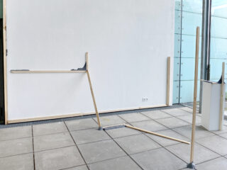 Mit grauem Material verbundene Holzstangen lehnen an einer weißen Wand und einem gefliesten Boden in einem minimalistischen, modernen Innenraum mit Glaspaneelen.