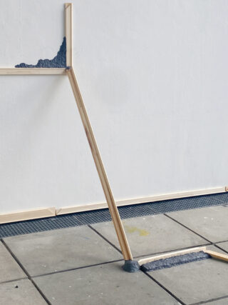 Ein minimalistischer Holzrahmen lehnt an einer weißen Wand, wobei Teile in raue Zementabschnitte an der Wand und dem gefliesten Boden eingebettet sind.