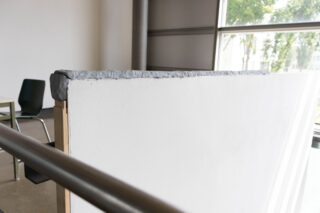 Eine Nahaufnahme einer weißen Trennwand mit grauem Kitt entlang der Oberkante, die in der Nähe eines Fensters in einem Raum mit einem Stuhl und einem Tisch im Hintergrund steht.