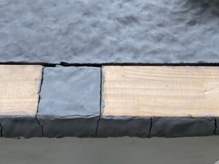 Eine Nahaufnahme eines Holzbretts, das teilweise mit einer Schicht aus grauem Ton bedeckt ist und sichtbare Risse und eine ungleichmäßige Textur aufweist.