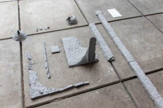 Graue, strukturierte Skulpturenobjekte in unterschiedlichen Formen und Größen sind geordnet auf einem gefliesten Betonboden angeordnet.