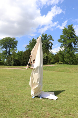 Eine Person steht auf Gras, ist größtenteils in ein großes Stück leichten Stoff gehüllt und hält eine Stange. Im Hintergrund sind Bäume und ein teilweise bewölkter Himmel zu sehen.
