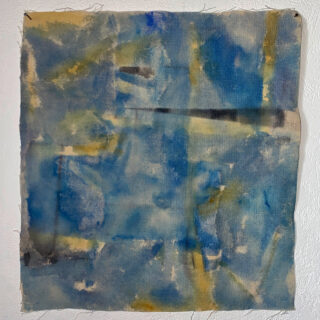 Ein quadratisches Stück Stoff mit abstrakten blauen, gelben und schwarzen Aquarellflecken und unebenen Rändern, an eine strukturierte weiße Wand gepinnt.