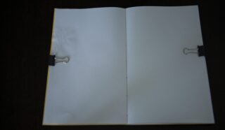 Ein offenes, leeres Notizbuch, das von zwei Heftklammern auf einer dunklen Oberfläche festgehalten wird.
