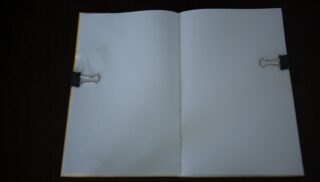 Ein offenes, leeres Notizbuch, das von zwei schwarzen Heftklammern auf einer dunklen Oberfläche festgehalten wird.