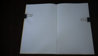 Ein offenes Notizbuch mit leeren weißen Seiten wird mit zwei schwarzen Heftklammern (eine auf jeder Seite) vor einem dunklen Hintergrund offen gehalten.