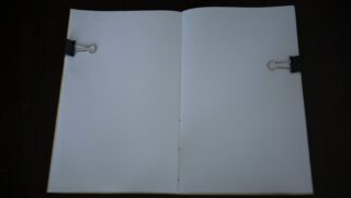 Ein offenes Notizbuch mit leeren weißen Seiten, die von zwei schwarzen Heftklammern (eine auf jeder Seite) flach gehalten werden, vor einem dunklen Hintergrund.