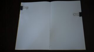 Ein offenes Heft mit leeren weißen Seiten, die von zwei schwarzen Heftklammern (eine auf jeder Seite) flach gehalten werden, vor einem dunklen Hintergrund.