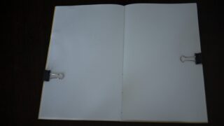Ein offenes, leeres Notizbuch wird von zwei schwarzen Heftklammern flach auf einer dunklen Oberfläche gehalten.