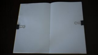 Ein offenes, leeres Notizbuch, das mit zwei Heftklammern (eine auf jeder Seite) vor einem dunklen Hintergrund gehalten wird.