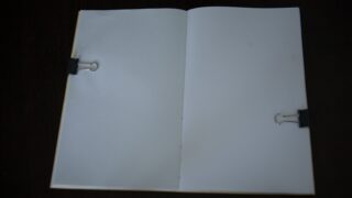 Ein offenes Heft mit leeren weißen Seiten wird links und rechts von schwarzen Heftklammern festgehalten.