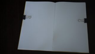 Ein offenes Notizbuch mit leeren weißen Seiten, die von zwei schwarzen Heftklammern (eine auf jeder Seite) vor einem dunklen Hintergrund festgehalten werden.