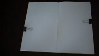 Ein offenes Notizbuch mit leeren weißen Seiten, das auf beiden Seiten von schwarzen Heftklammern gehalten wird, liegt auf einer dunklen Oberfläche.
