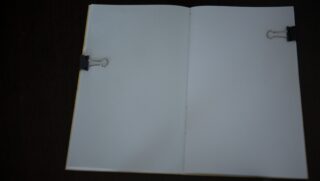 Vor einem dunklen Hintergrund steht ein offenes Notizbuch mit leeren weißen Seiten, das von zwei schwarzen Heftklammern auf gegenüberliegenden Seiten flach gehalten wird.