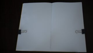Ein offenes, leeres Notizbuch, das an den Seiten mit zwei schwarzen Heftklammern gehalten wird und auf einer dunklen Oberfläche liegt.