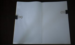 Ein offenes Notizbuch mit leeren weißen Seiten, die von zwei schwarzen Heftklammern (eine auf jeder Seite) auf einer dunklen Oberfläche festgehalten werden.