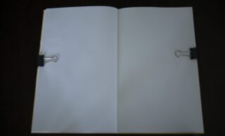 Ein offenes Notizbuch mit leeren weißen Seiten, die mit schwarzen Heftklammern auf einer dunklen Oberfläche festgehalten werden.