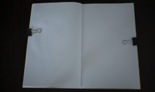 Ein offenes, leeres Notizbuch, flach gehalten mit zwei schwarzen Heftklammern, eine auf jeder Seite, vor einem dunklen Hintergrund.