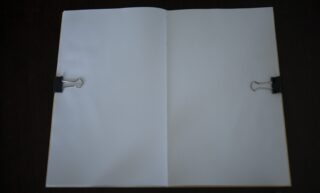 Ein offenes Notizbuch mit leeren weißen Seiten, die auf beiden Seiten mit schwarzen Heftklammern festgehalten werden, liegt auf einer dunklen Oberfläche.