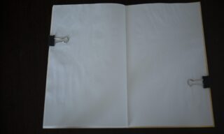Ein offenes, leeres Notizbuch wird mit zwei schwarzen Heftklammern flach auf einer dunklen Oberfläche gehalten.