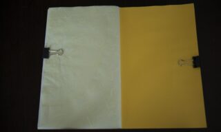 Auf einer dunklen Oberfläche liegt ein geöffneter Ordner mit einer weißen strukturierten linken Seite und einer gelben rechten Seite, die jeweils mit einer schwarzen Büroklammer festgehalten werden.