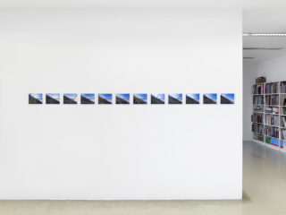 Eine Reihe kleiner Fotografien ist in gleichmäßigen Abständen in einer horizontalen Linie an einer weißen Galeriewand angeordnet. Im Hintergrund rechts sind Bücherregale zu sehen.