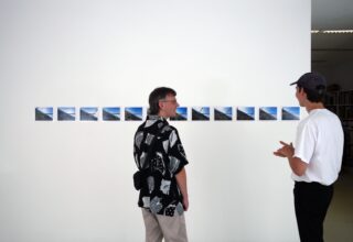 Vor einer weißen Wand, an der horizontal eine Reihe kleiner Landschaftsfotos angebracht sind, stehen zwei Personen und unterhalten sich.