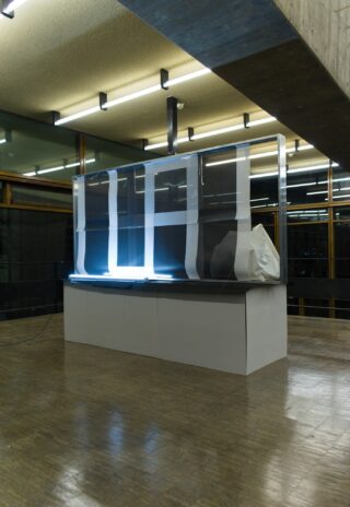 Eine große rechteckige Struktur mit transparenten Platten, weißem Klebeband und einem in Plastik eingewickelten Objekt steht in einem modernen, mit Leuchtstoffröhren beleuchteten Raum mit Betonböden und Glaswänden.
