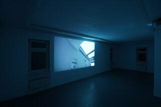 Ein dunkler Raum mit einer großen Wandprojektion, die das Bild einer Decke, eines Oberlichts und eines Heizkörpers zeigt, die von einem blauen Farbton beleuchtet werden.