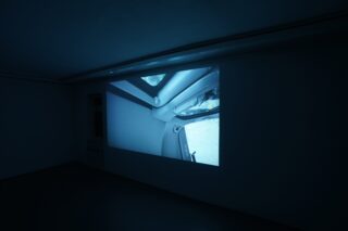 Eine Videoprojektion auf einer dunklen Wand zeigt eine Innenansicht mit einem Fenster und Rohren, möglicherweise aus dem Inneren eines Fahrzeugs oder einer Industrieanlage.
