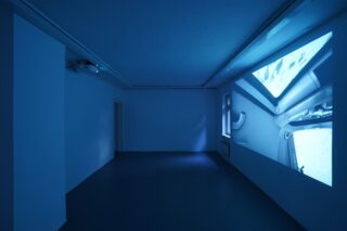 Ein dunkler, leerer Raum mit blauer Beleuchtung und einer großen Videoprojektion an der Wand, die einen Teilblick in das Innere eines Raumschiffs zeigt.