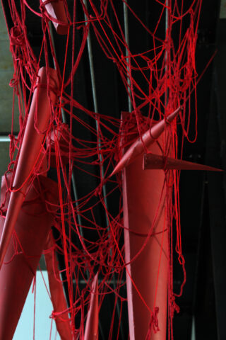 Vor einem dunklen Industriehintergrund sind rote Seile um mehrere vertikal aufgehängte rote Metallrohre und -stangen gewickelt.