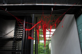 Eine rote Fadenkunstinstallation, die einem Netz ähnelt, ist zwischen Wänden und Metallträgern in einem Innenkorridor aufgehängt, im Hintergrund befinden sich Treppen und Fenster.