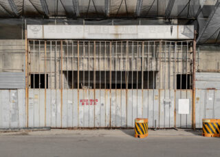 Ein geschlossenes Metalltor mit verrosteten Gitterstäben und verblasster koreanischer Beschilderung darüber, zwei gelb gestreifte Verkehrszeichen davor und ein Warnschild am Tor.