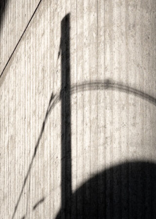 Der Schatten einer Straßenlaterne und ihres gebogenen Arms wird auf eine strukturierte, vertikal gerillte Betonwand geworfen.