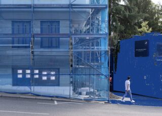 Eine Person geht an einem im Bau befindlichen Gebäude vorbei, das mit einem blauen Netz bedeckt ist. In der Nähe befindet sich eine blaue temporäre Struktur und im Hintergrund sind Bäume zu sehen.
