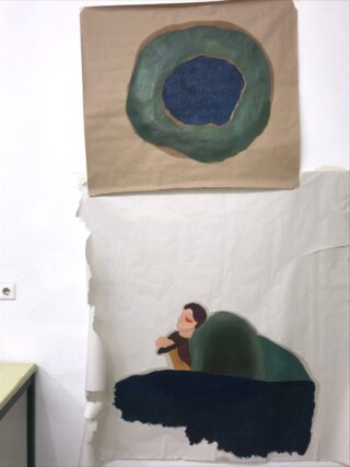 An einer weißen Wand hängen zwei abstrakte Gemälde; das obere zeigt eine grüne kreisförmige Form, während das untere eine Person zeigt, die neben einer großen grünen Form sitzt.