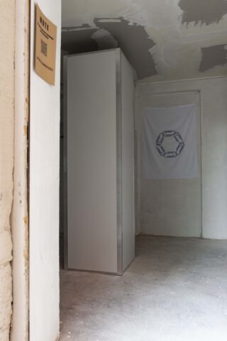 In einem kahlen Raum mit unfertigen Wänden steht eine hohe, schlichte rechteckige Struktur; an der Rückwand hängt eine weiße Flagge mit einem kreisförmigen Kettenmuster.